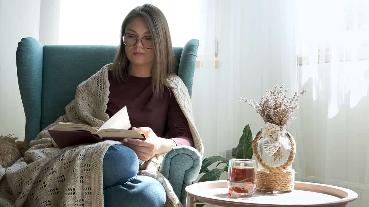 年轻女子戴眼镜看书，覆盖舒适的格子，坐在家里的扶手椅视频素材