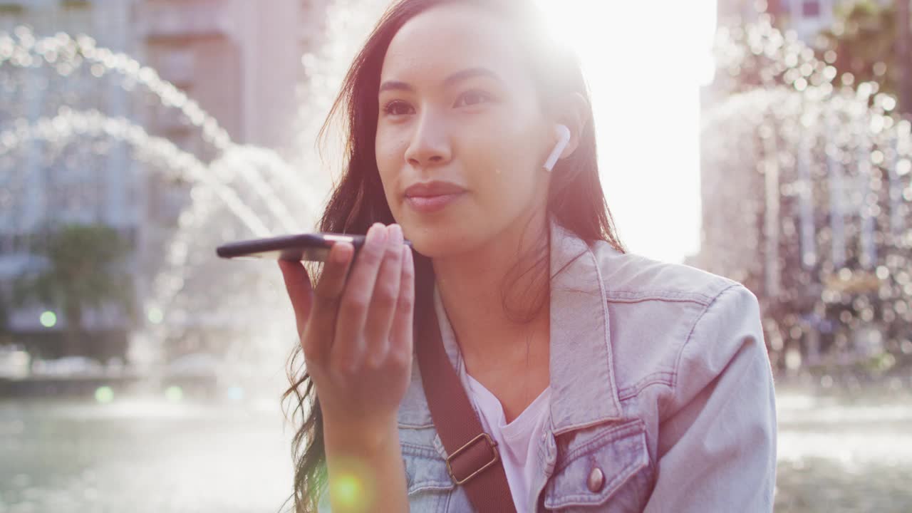 一名亚洲女子坐在喷泉旁，戴着耳机，用智能手机聊天视频素材