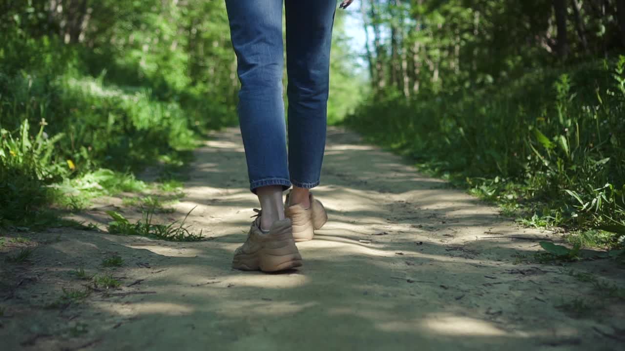 年轻时髦的女人走在夏日森林小径Spbd上。穿着牛仔裤和运动鞋的女孩的旅程视频素材