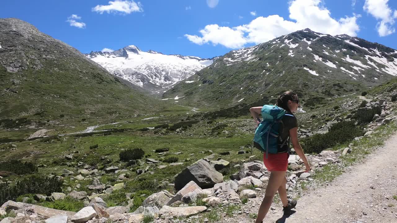 一名身穿短裤的女子在奥地利卡林西亚的安科格尔徒步旅行。那女人走在一条砾石路上。后面是高冰川。四周牧场绿油油。阿尔卑斯山的早春。宁静视频素材