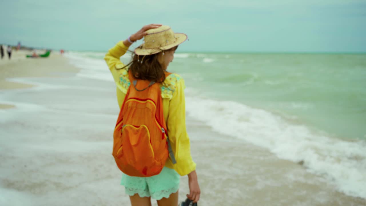 梦想自由的女人穿着夏天鲜艳的衣服，背着小背包在海边散步，在海滩上享受暴风雨和大风的天气，玩海浪视频素材