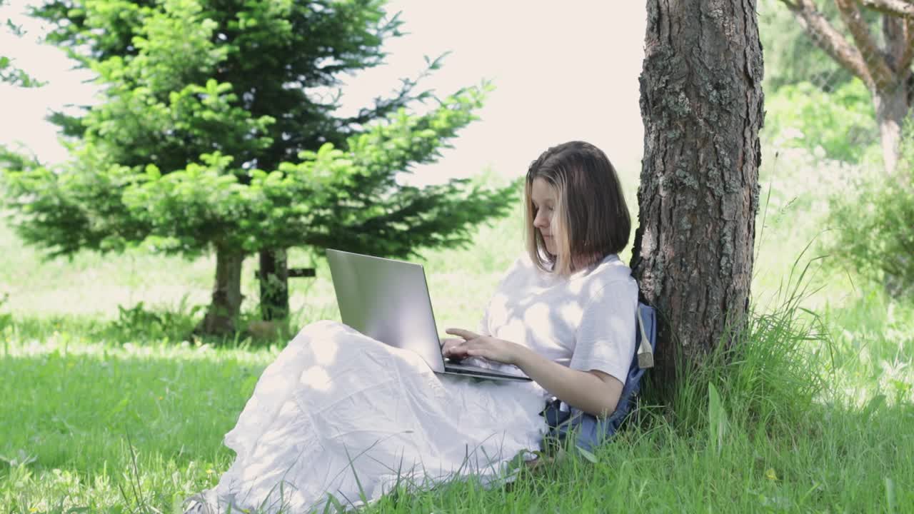 少女坐在公园树旁的草地上，在笔记本电脑上打印。年轻女子抬起头，微笑着挥了挥手视频素材