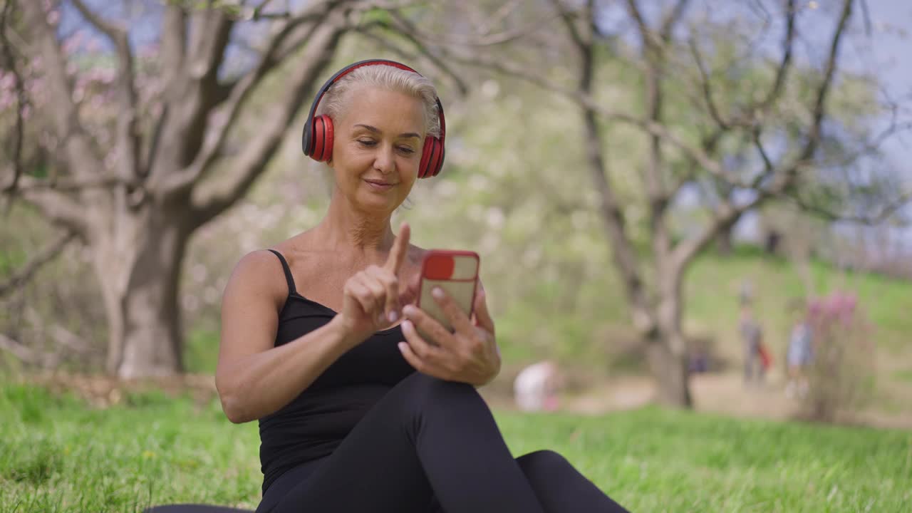 迷人的健身女运动员打开智能手机上的音乐在慢动作坐在阳光明媚的夏天公园。积极快乐的白人退休享受爱好和健康的生活方式视频素材