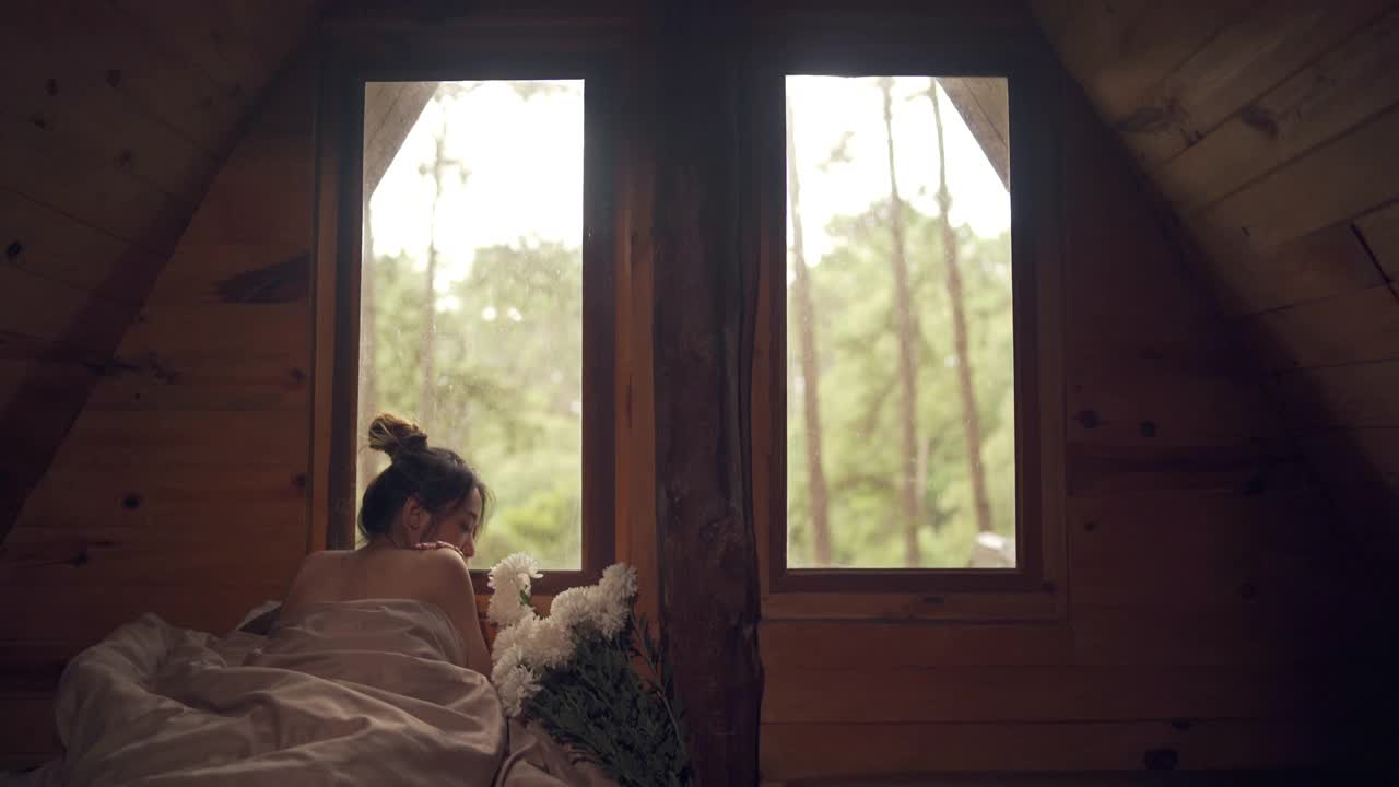 迷人的亚洲女人躺在床上的窗户与看美丽的自然外木屋在秋天的早晨。微笑漂亮的女孩放松，享受假期休息。视频素材