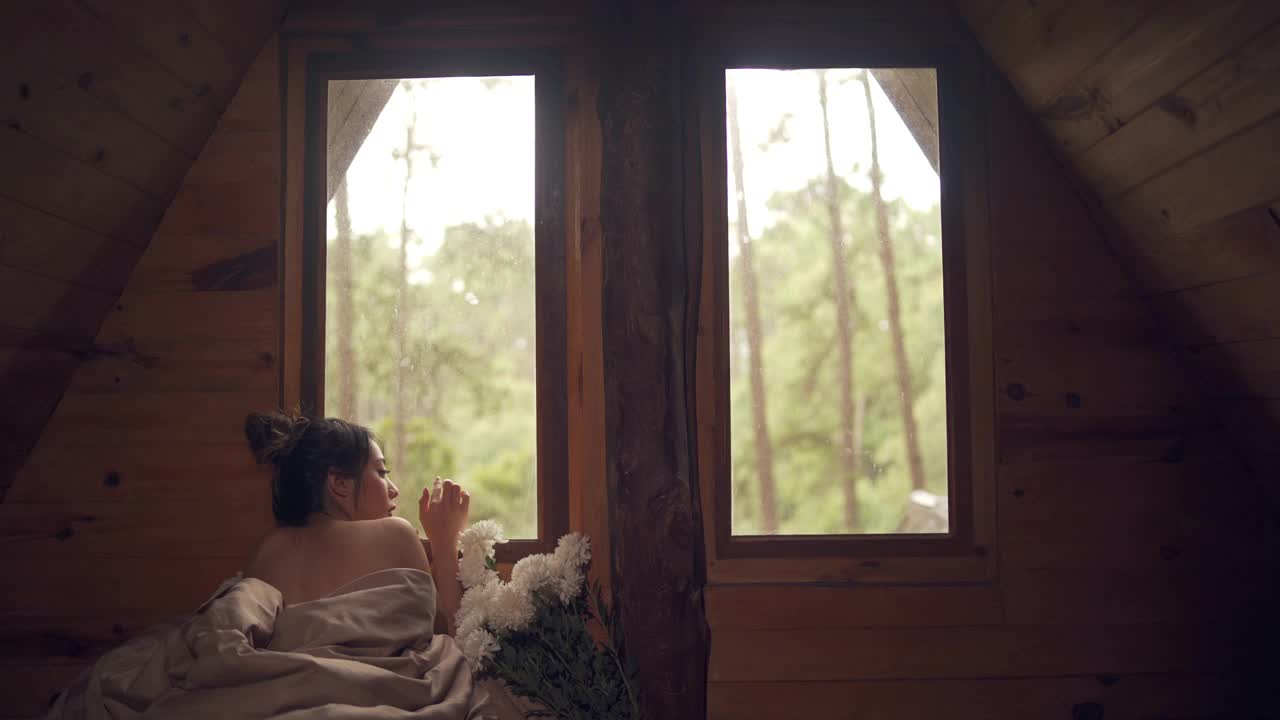 迷人的亚洲女人躺在床上的窗户与看美丽的自然外木屋在秋天的早晨。微笑漂亮的女孩放松，享受假期休息。视频素材