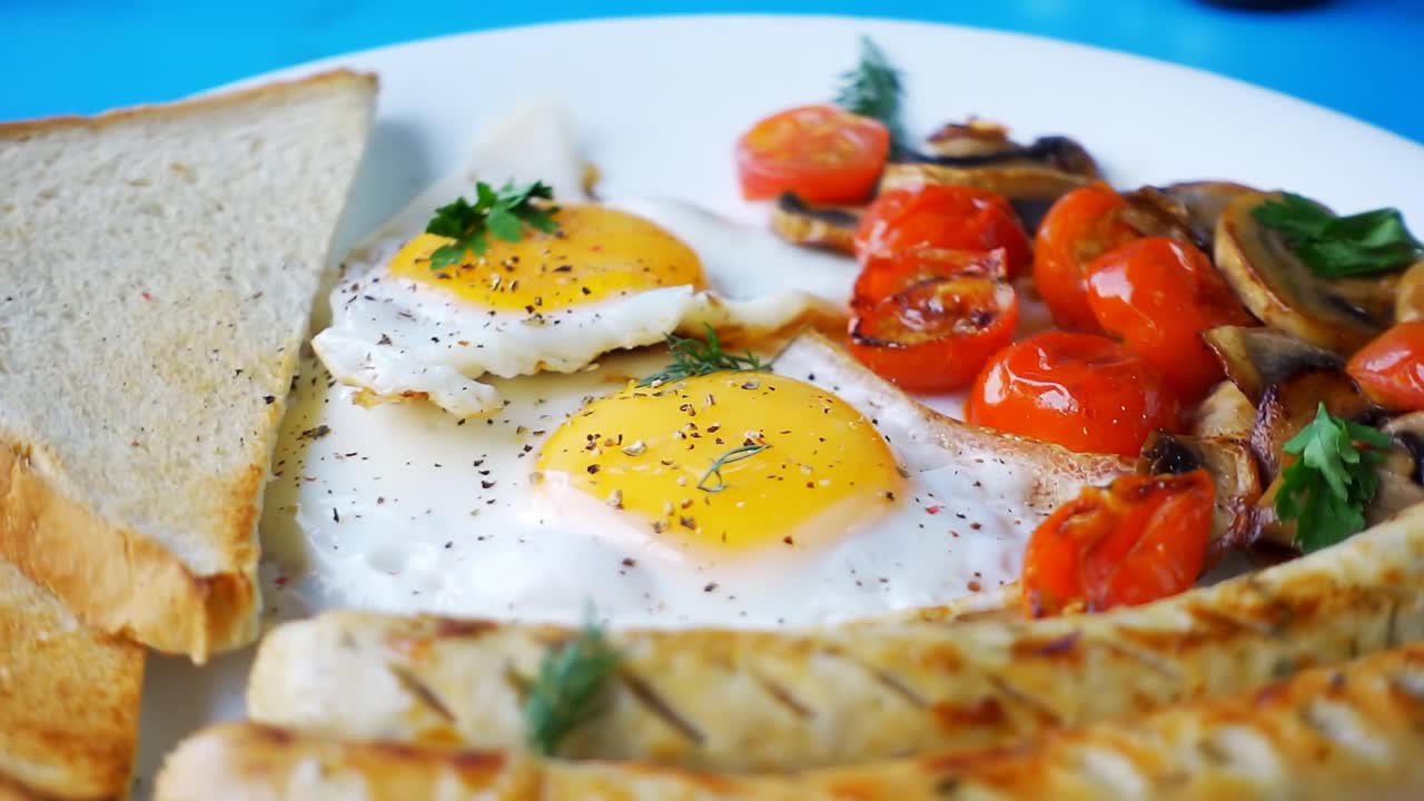 慢动作视频中香料落在煎鸡蛋的蛋黄上，在蓝色背景的早餐盘上的特写镜头。早餐在白色的盘子里旋转。视频素材