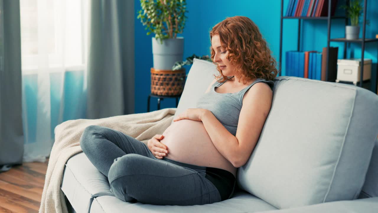 一个美丽迷人的年轻女子双腿交叉坐在沙发上，和她怀孕的肚子聊天，抚摸它，触摸，讲婴儿的故事，等不及要生产，温柔的微笑，深情的回头看视频素材