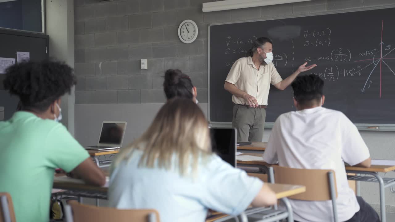 数学老师和学生在教室里戴着防护口罩。数学公式写在黑板上的高中学院。冠状病毒大流行中的教育和医疗视频素材