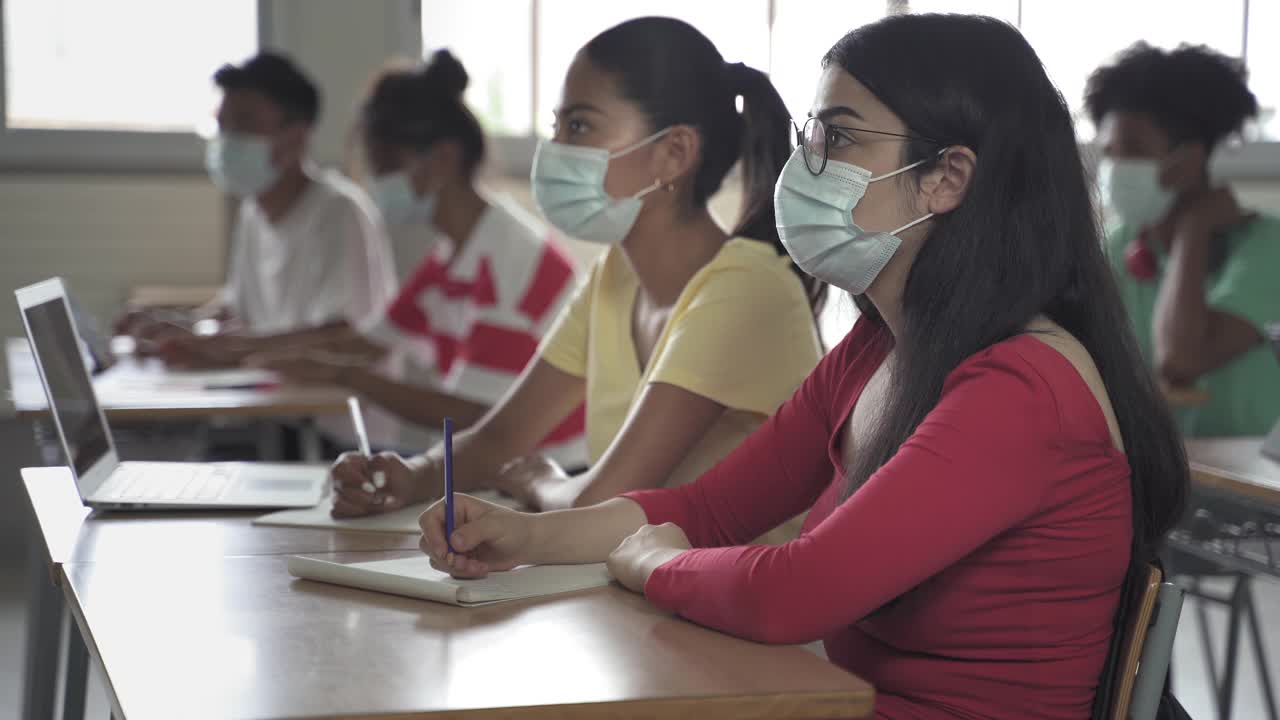 中学生戴着医用口罩在高中或大学课堂上写笔记。新冠肺炎疫情下的返校视频素材