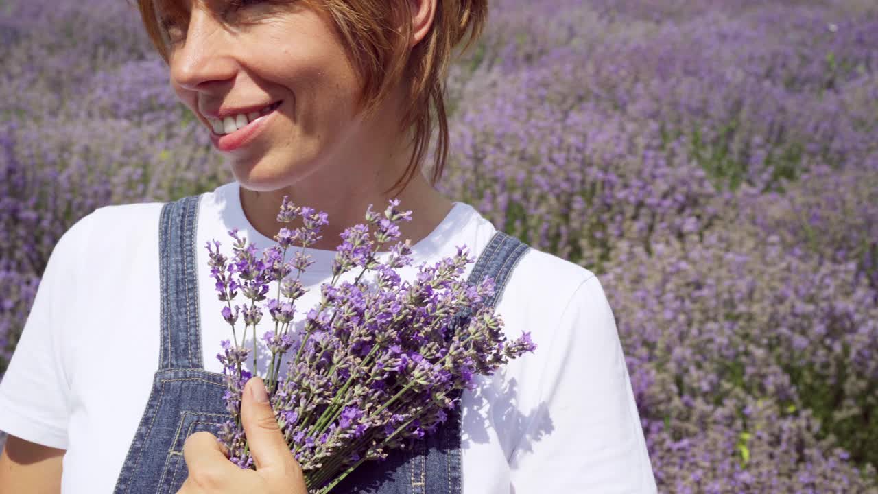 迷人的微笑的女人拿着花束站在薰衣草田。白人女士享受阳光灿烂的夏天在鲜花草地上。幸福和女性的概念。视频素材