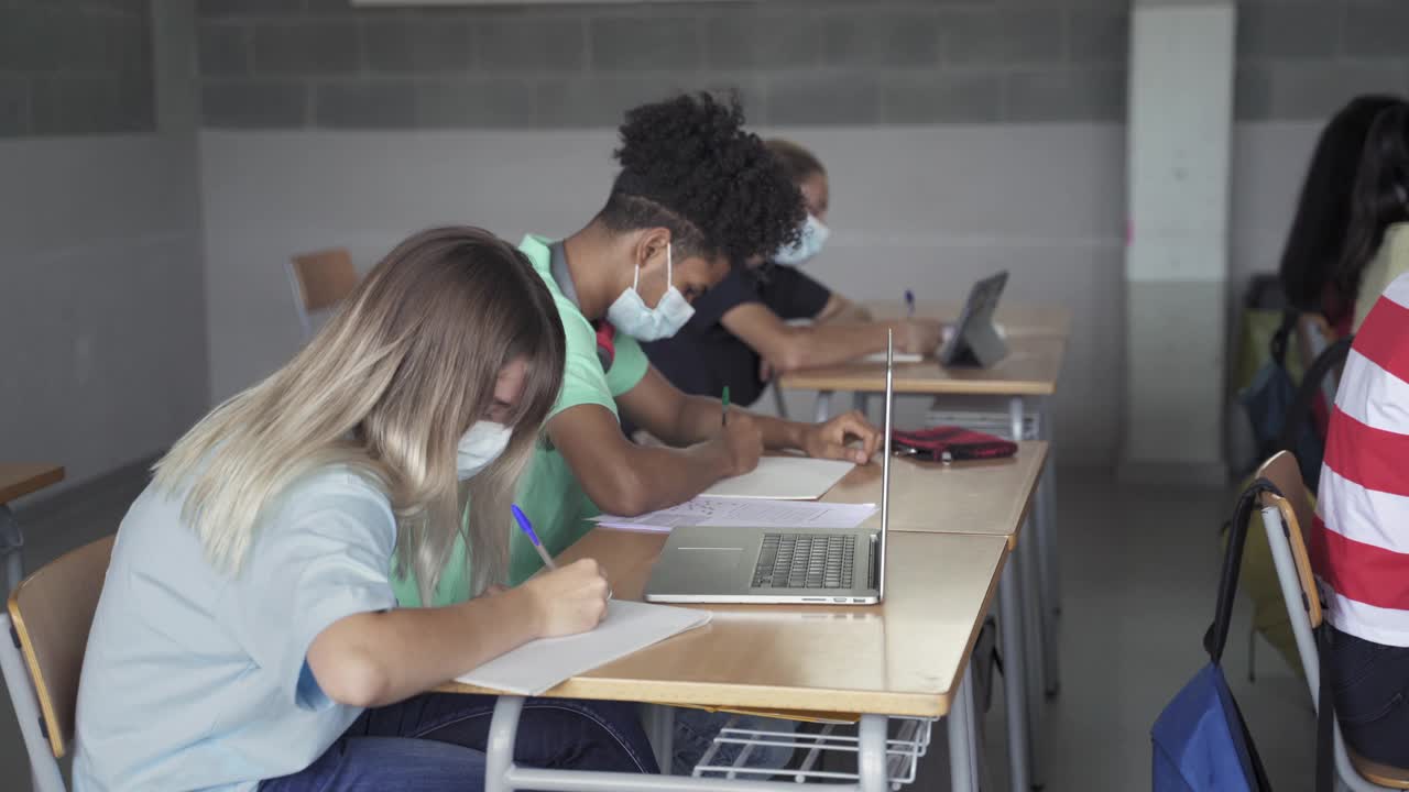 戴着防护口罩的中学生在高中课堂上写笔记。冠状病毒大流行中的教育和医疗视频素材