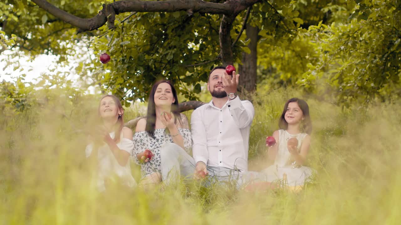 父母带着孩子向空中扔苹果。快乐的父母和两个可爱的女儿坐在绿色花园的格子毯子上吃苹果。一个白人家庭在新鲜空气下进行夏季野餐视频素材