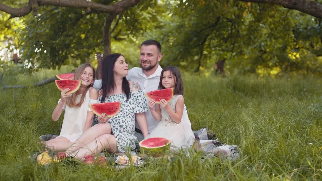 一对白种人夫妇和两个小女儿紧挨着坐在绿色的花园里，手里拿着几块西瓜。积极的家庭享受一起野餐的时间。家庭和孩子们视频素材