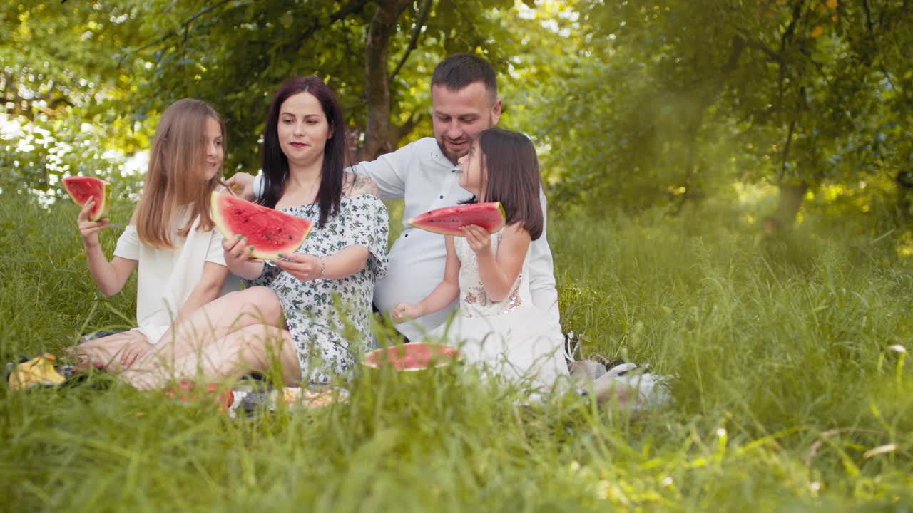 兴高采烈的父母带着女儿们，穿着夏装，坐在绿色的花园里，享受着甜甜的西瓜。在户外野餐。弛豫时间。父母带着孩子野餐，带着甜甜的水果视频素材