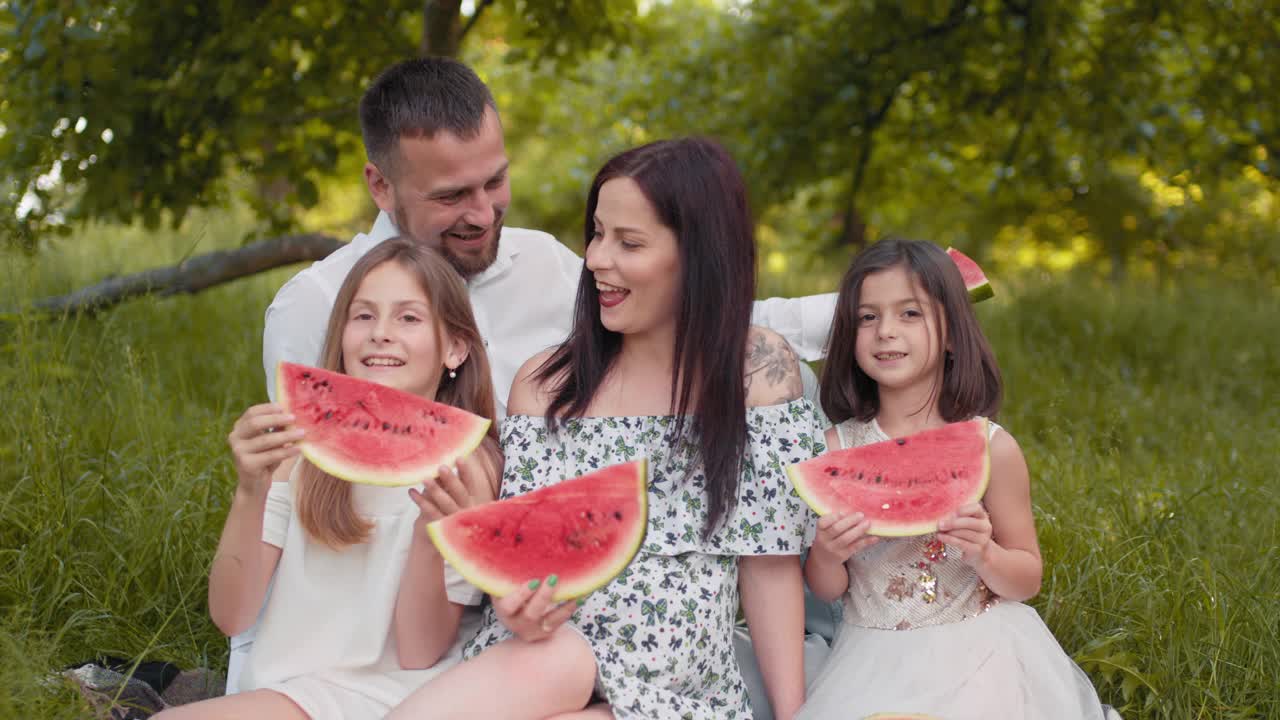 快乐的家庭和孩子们在绿色公园夏天野餐。年轻的父母和两个女儿在吃甜西瓜。户外放松和享受。父母带着两个孩子在野餐时吃西瓜视频素材