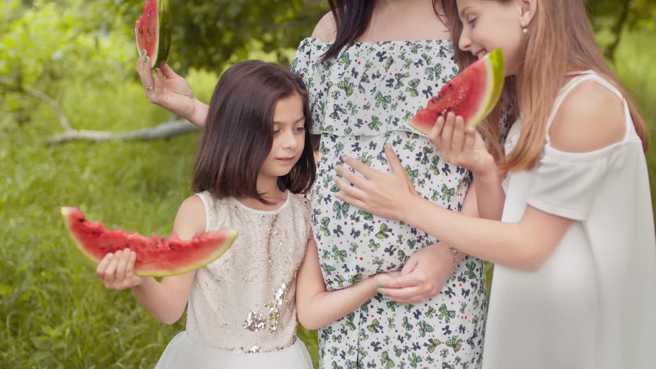 怀孕的白人妇女和她的两个漂亮的女儿站在一起在绿色花园拥抱。幸福家庭在户外吃甜西瓜。周末时间。女人和孩子们在户外吃西瓜视频素材