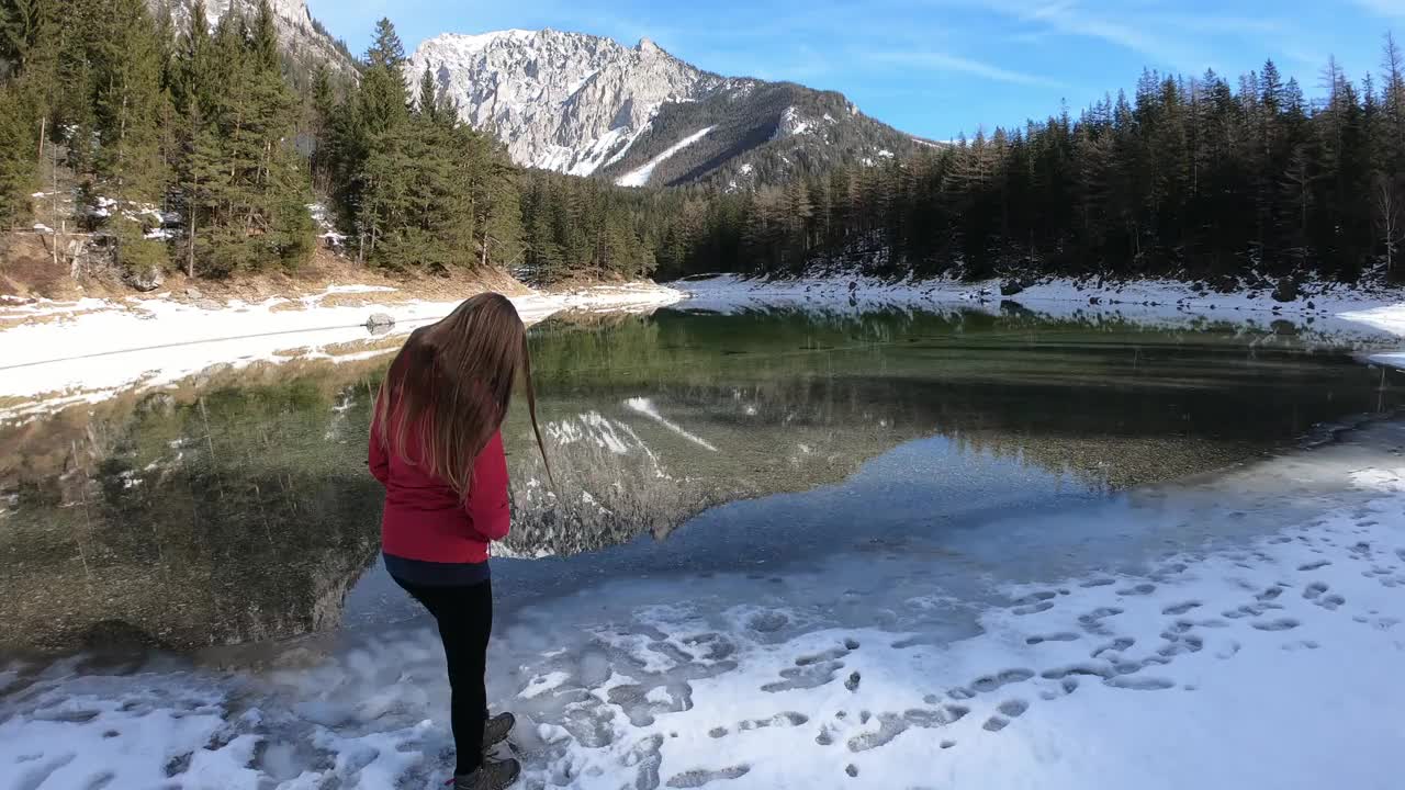 冬天，一个穿着粉红色夹克的女人在奥地利的绿湖附近散步。湖被雪包围着。湖的四周有一片茂密的森林。清水。阳光明媚的一天。放松视频素材