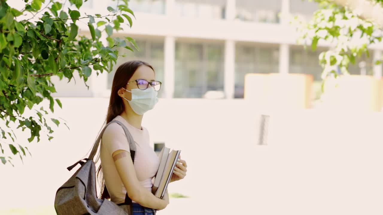 新冠肺炎疫情结束后，接种疫苗的学生返回学校或大学，一名少女背着背包和书籍站在学校旁边，手上拿着膏药视频素材