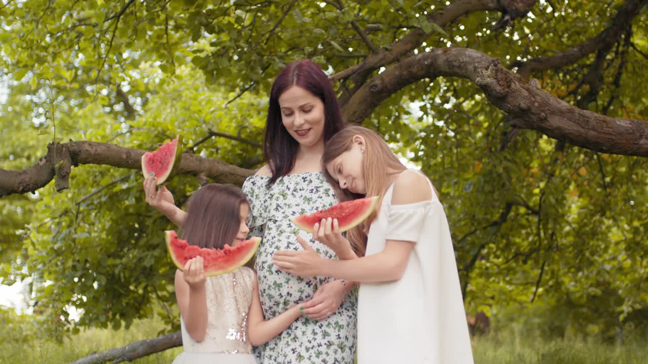 怀孕的白人妇女和她的两个漂亮的女儿站在一起在绿色花园拥抱。幸福家庭在户外吃甜西瓜。两个女孩在花园里抚摸着怀孕母亲的肚子视频素材