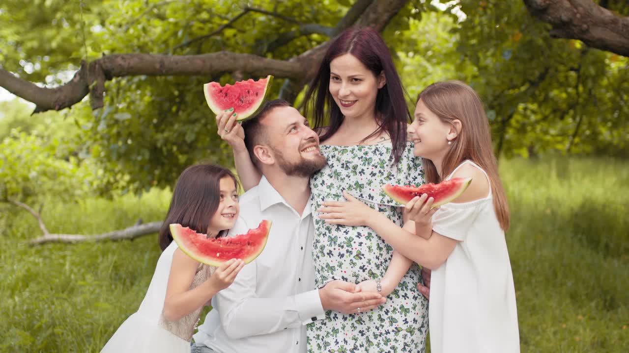 正白种人一家人穿着夏装一起在绿园摆造型，吃着甜西瓜。帅哥和两个可爱的孩子拥抱孕妇。带着丈夫和孩子在户外的孕妇视频素材