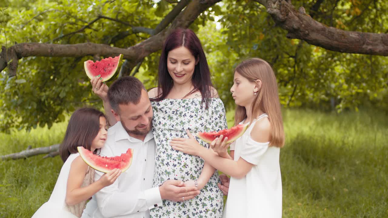 正白种人一家人穿着夏装一起在绿园摆造型，吃着甜西瓜。帅哥和两个可爱的孩子拥抱孕妇。有两个孩子的孕妇视频素材