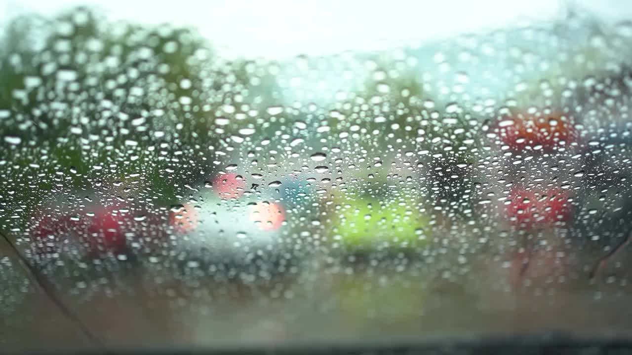 靠近汽车镜子上的雨滴。开车时，雨点落在汽车挡风玻璃上。街道外车辆的彩色模糊的灯光。视频素材