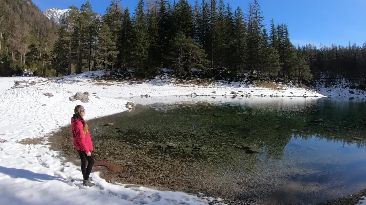 冬天，一个穿着粉红色夹克的女人在奥地利的绿湖附近散步。湖被雪包围着。湖的四周有一片茂密的森林。清水。阳光明媚的一天。放松视频素材