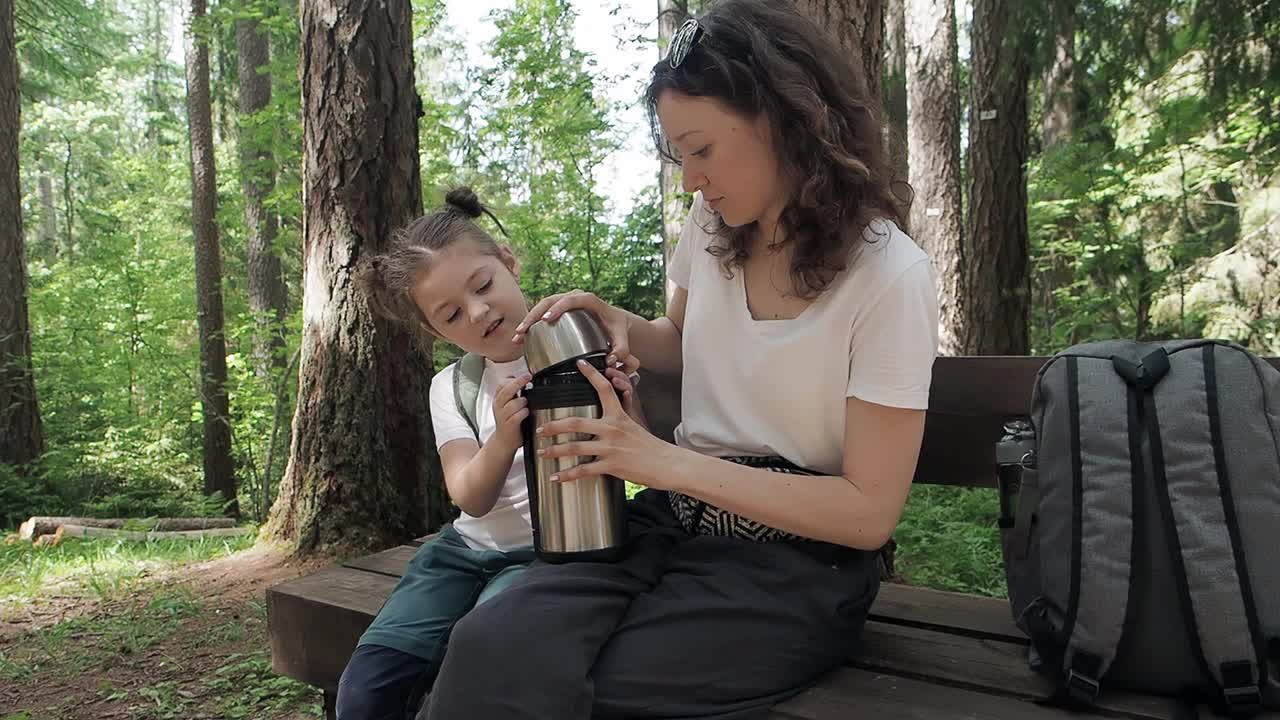 旅行者的母亲和女儿打开热茶保温瓶坐在长凳上在森林夏天远足。幸福的家庭在假期旅行中享受清爽和放松的时刻。质量的时间在一起视频下载