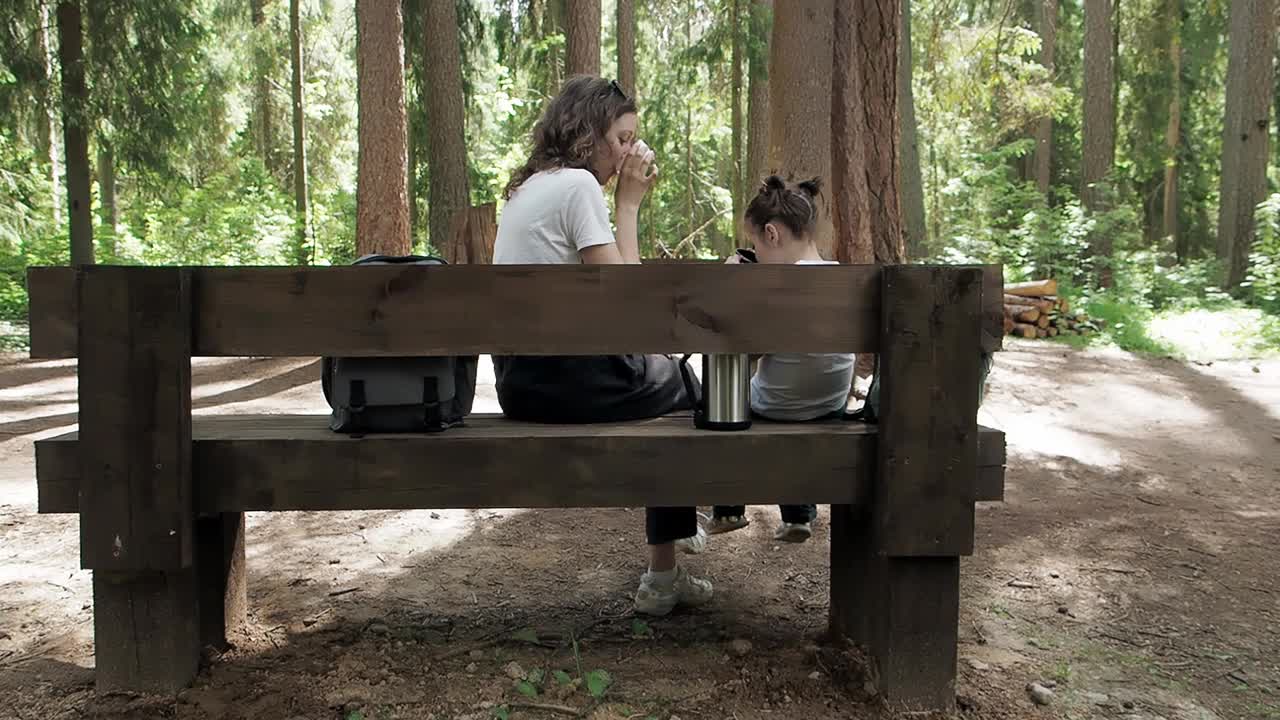 后视图。旅行者女人和女孩喝热茶从保温杯坐在长凳上在森林夏天远足。幸福家庭享受热饮。在假期旅行中放松身心视频下载