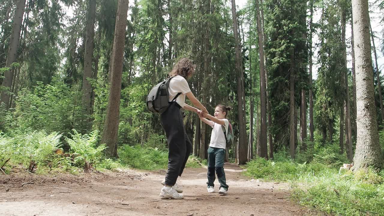 快乐微笑的女孩紧紧抓住她年轻的母亲的手环绕在夏天绿色的森林公园小径。母女时间在一起度假没有蚊子，家庭时间概念。慢动作视频素材