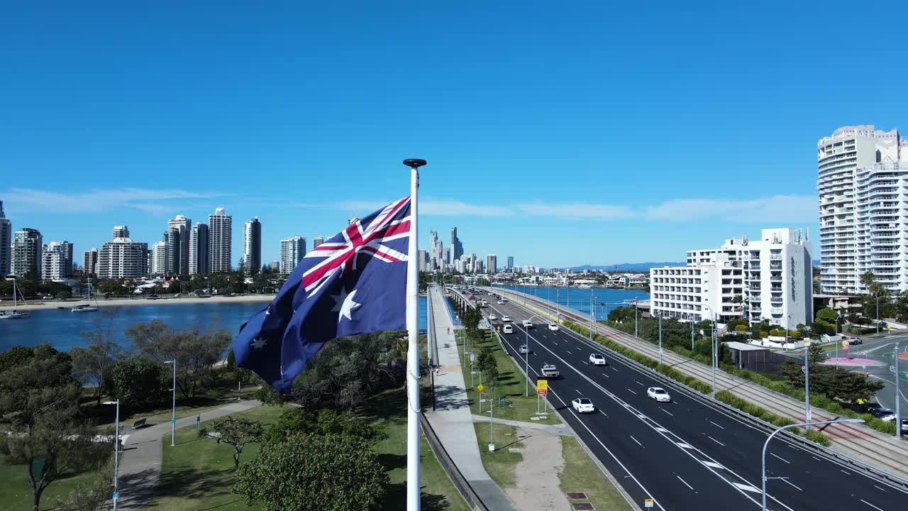 近距离观察澳大利亚国旗在强风中飘扬，在繁忙的道路网络下，通往高耸的城市天际线。视频素材