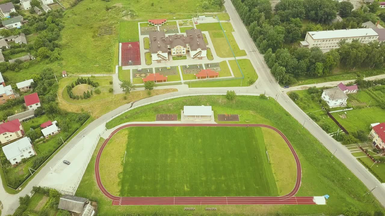 农村学校建筑附近的体育场鸟瞰图，红色的跑道和绿色的草地足球场。视频素材