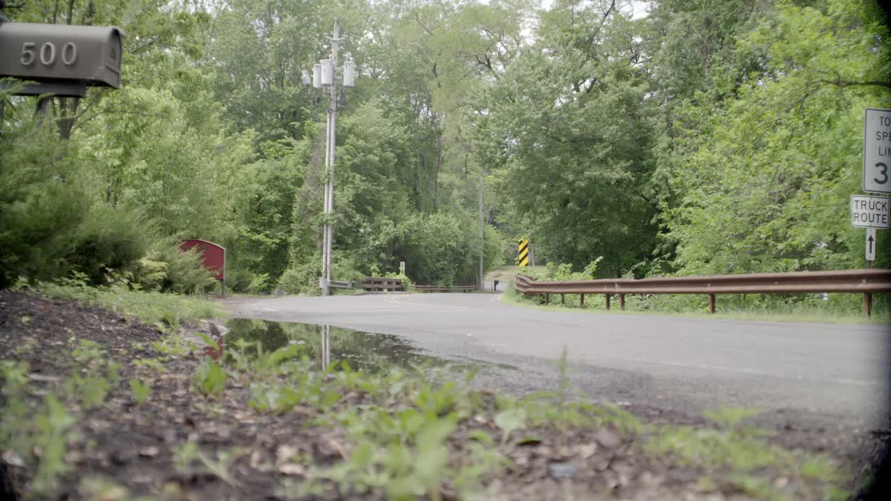 乡村道路弯角大。可见树木和限速标志。邮箱在fg中可见。在bg中可见的电话杆。可能是郊区。从bg开车到fg。汽车撞到水坑里溅起了水花。视频素材