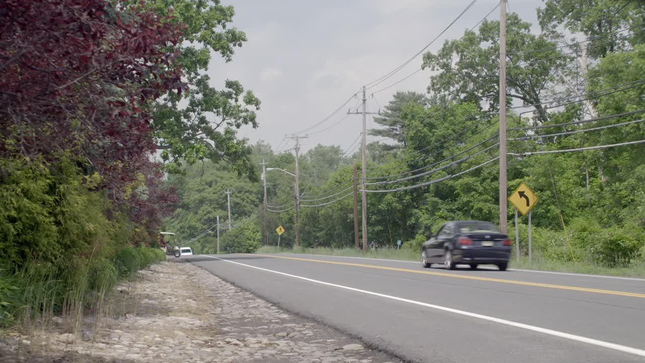 广角的乡村道路两旁有树木和电线杆。可见骑自行车的人或骑自行车的人。汽车在路上朝两个方向行驶。可能是郊区。高尔夫球车开过马路。视频素材