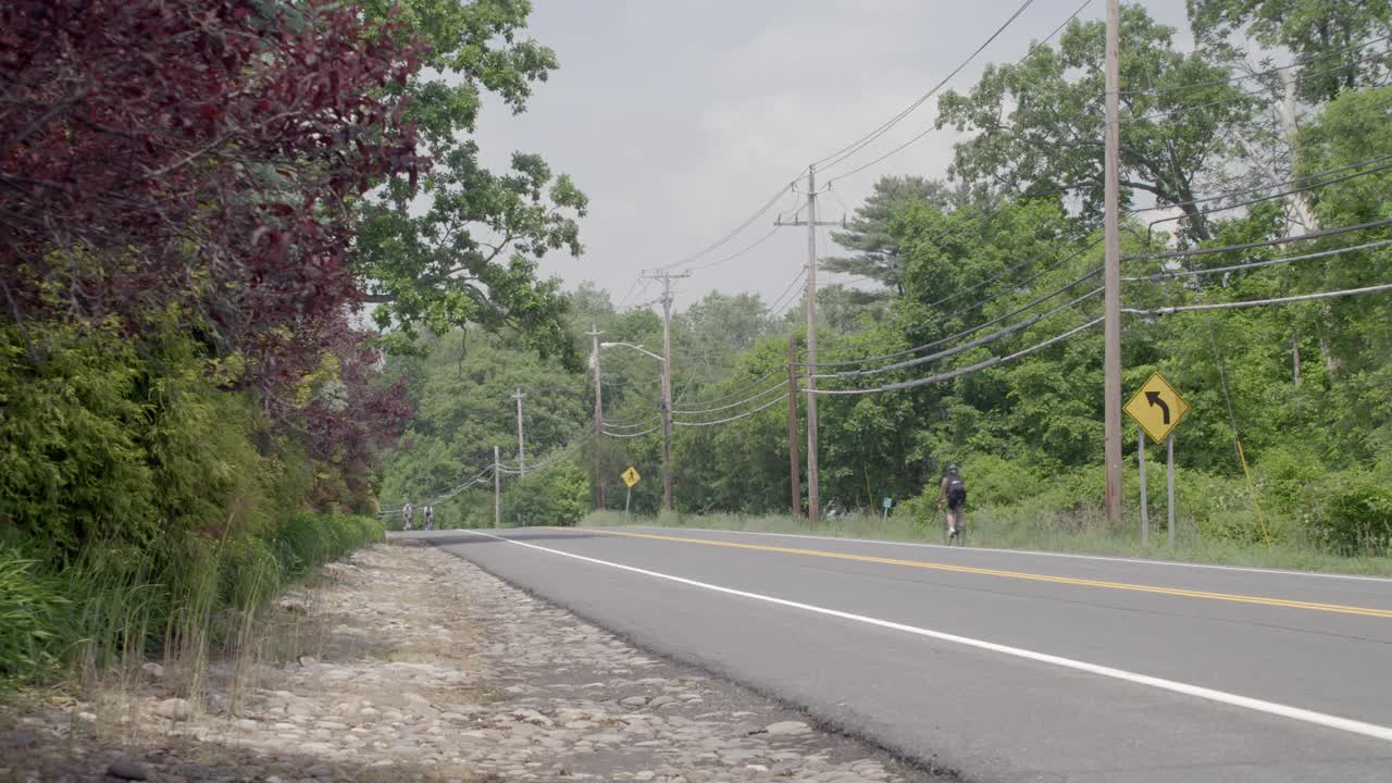 广角的乡村道路两旁有树木和电线杆。可见骑自行车的人或骑自行车的人。汽车在路上朝两个方向行驶。可能是郊区。视频素材