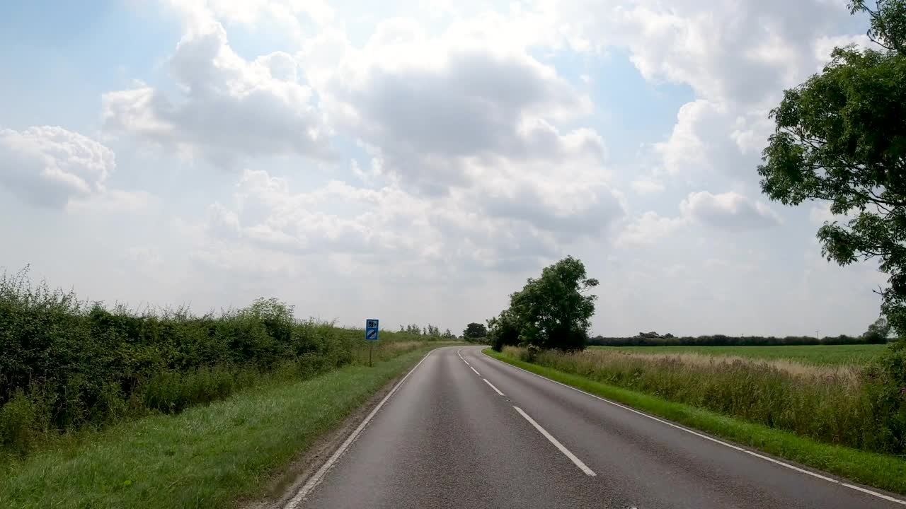 4K实时POV汽车行驶在英国的乡村公路长直。农场，田野和树篱路在一个阳光灿烂的蓝天。非城市场景的观点视频素材