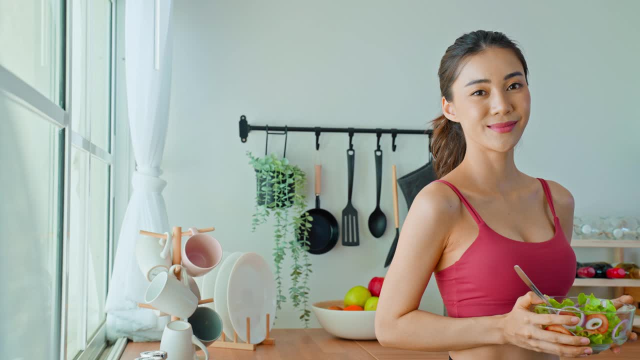 亚洲美女肖像拿着沙拉碗看着镜头。漂亮的运动女孩穿着运动服在家里享受运动后吃干净的蔬菜的健康。饮食和健康食品的概念。视频素材