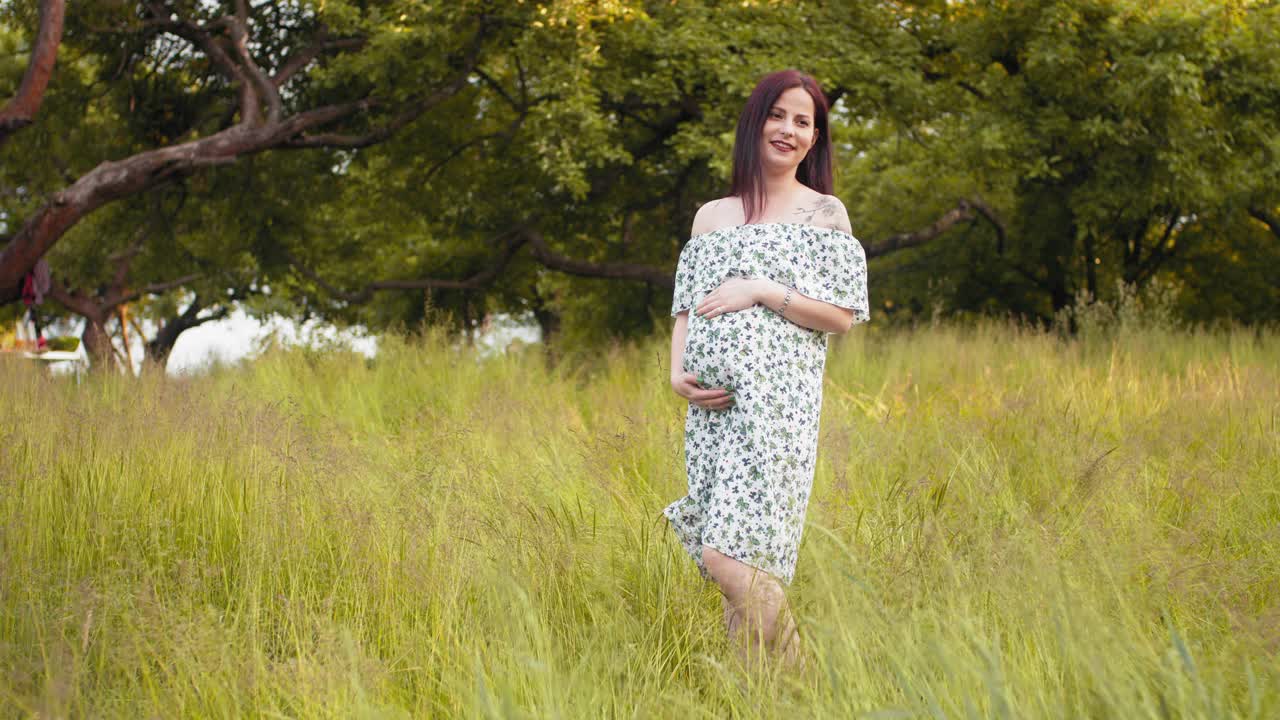 漂亮的高加索女人在夏天的裙子爱抚她怀孕的肚子，而在绿色的花园。期望观念与健康妊娠。迷人的孕妇穿着夏装在户外摆姿势视频素材