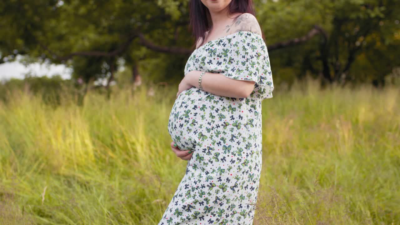 年轻的女士双手放在她怀孕的肚子上，站在绿色的花园。夏天在大自然中放松。幸福的概念。孕妇穿着衣服在户外摆姿势视频素材