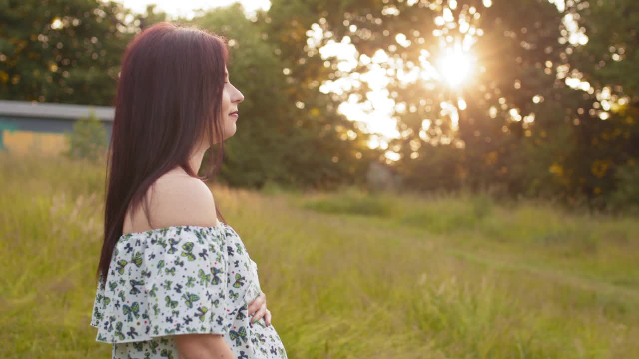 可爱的黑发年轻女子站在绿色花园，抱着她怀孕的肚子。快乐的女士穿着时尚的夏装和裸露的肩膀。夏装女子站在绿色的花园视频素材