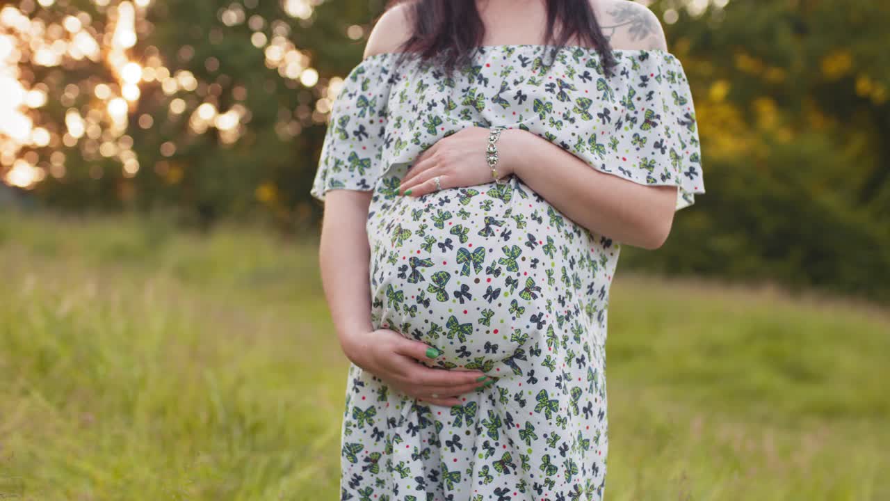 一名身着夏装的白人孕妇在户外摆姿势。年轻的女士双手放在她怀孕的肚子上，站在绿色的花园。夏天在大自然中放松视频素材