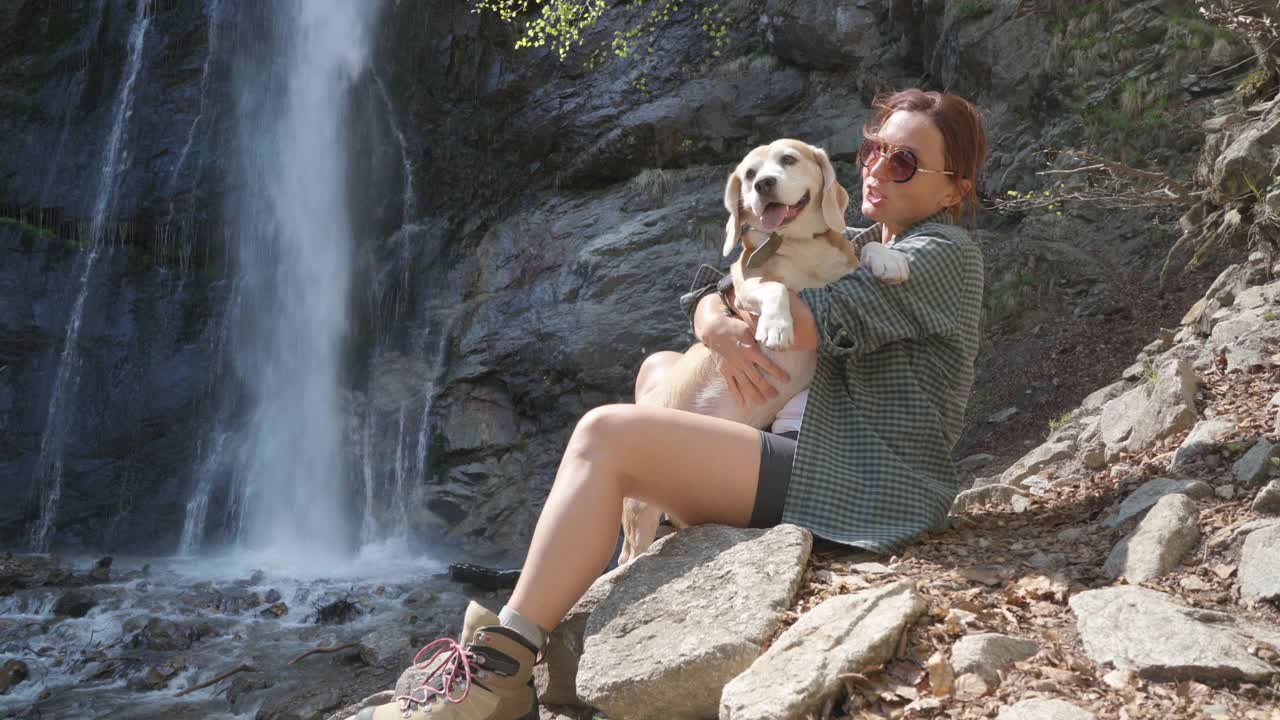 一个女人坐在瀑布附近，抱着她的小猎犬视频素材