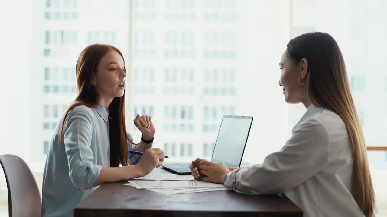 两名同事的侧视图，年轻的商业女性员工在工作场所一起坐在书桌上的窗口背景与城市景观。视频素材