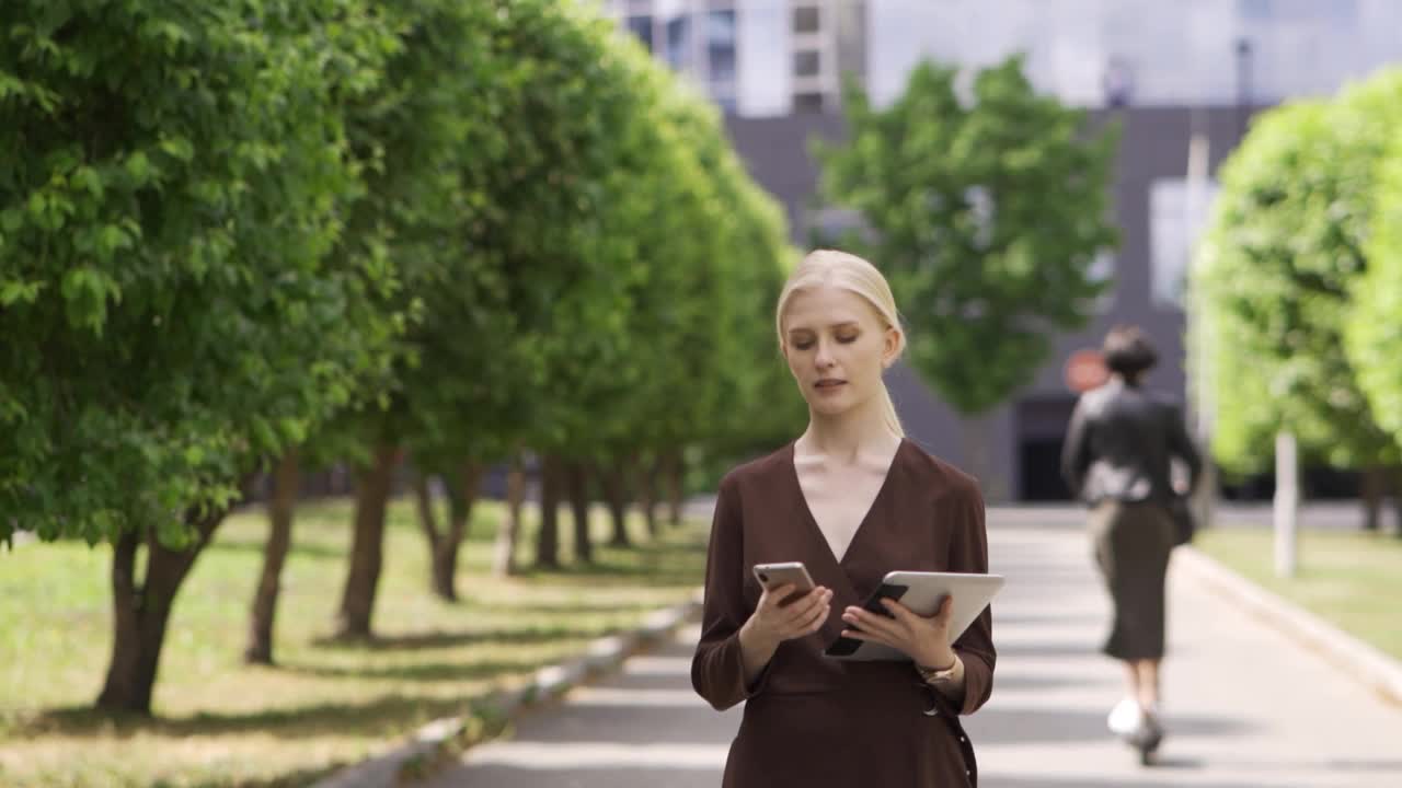 一个年轻的女商人走在街上，手里拿着平板电脑，正在讲电话。在她的身后是高楼大厦和一排排种植的树木。视频素材