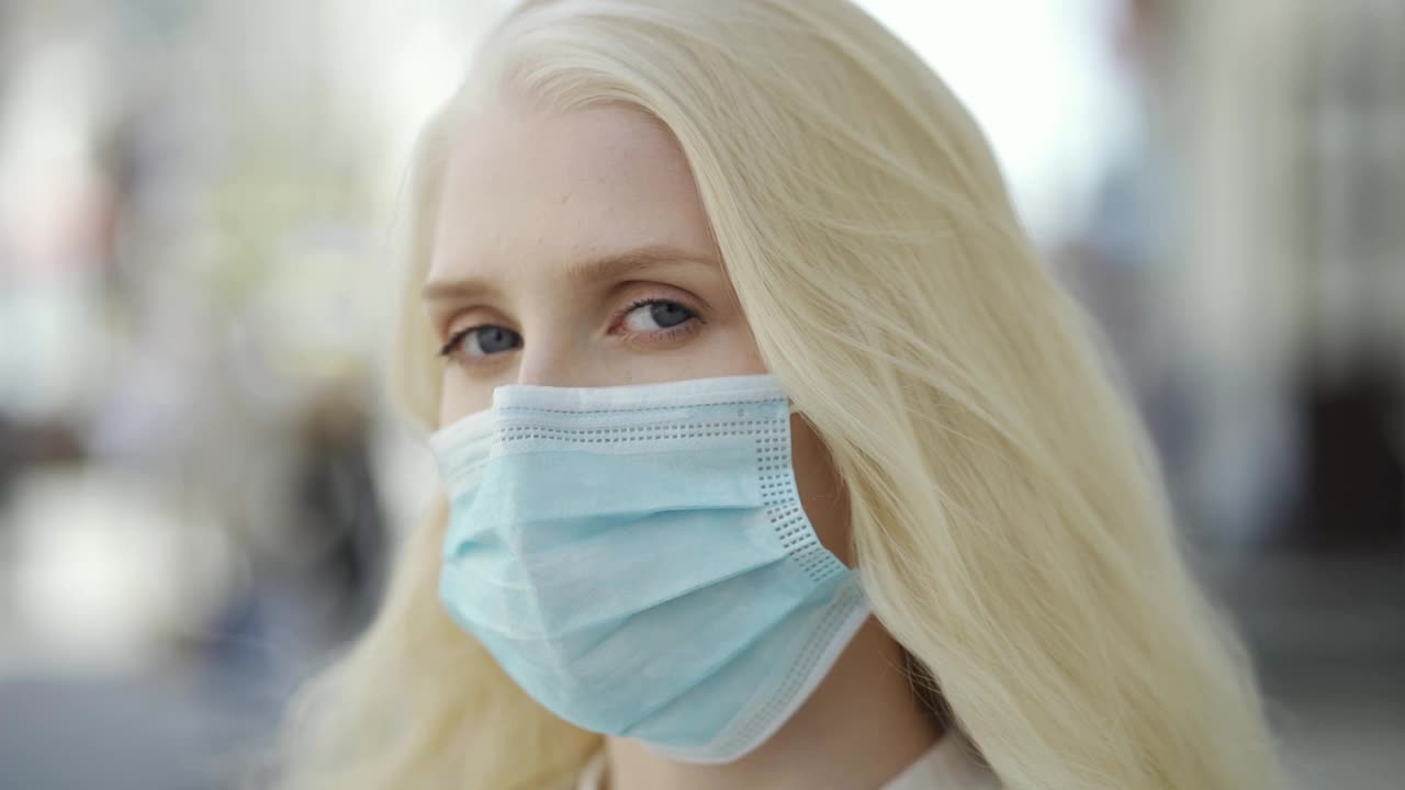 一个戴着医用口罩的蓝眼睛金发女人站在一个大城市的中心街道上看着摄像机。冠状病毒疫情、个人防护装备、检疫隔离。视频购买
