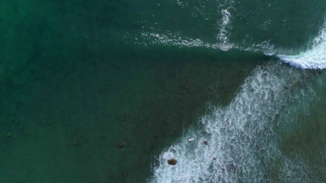 海洋表面的垂直视图。在长板上冲浪者向波浪移动。冲浪者试图克服来自海浪的泡沫。在海面下可以看到暗礁和岩石。冲浪者的俯视图视频素材