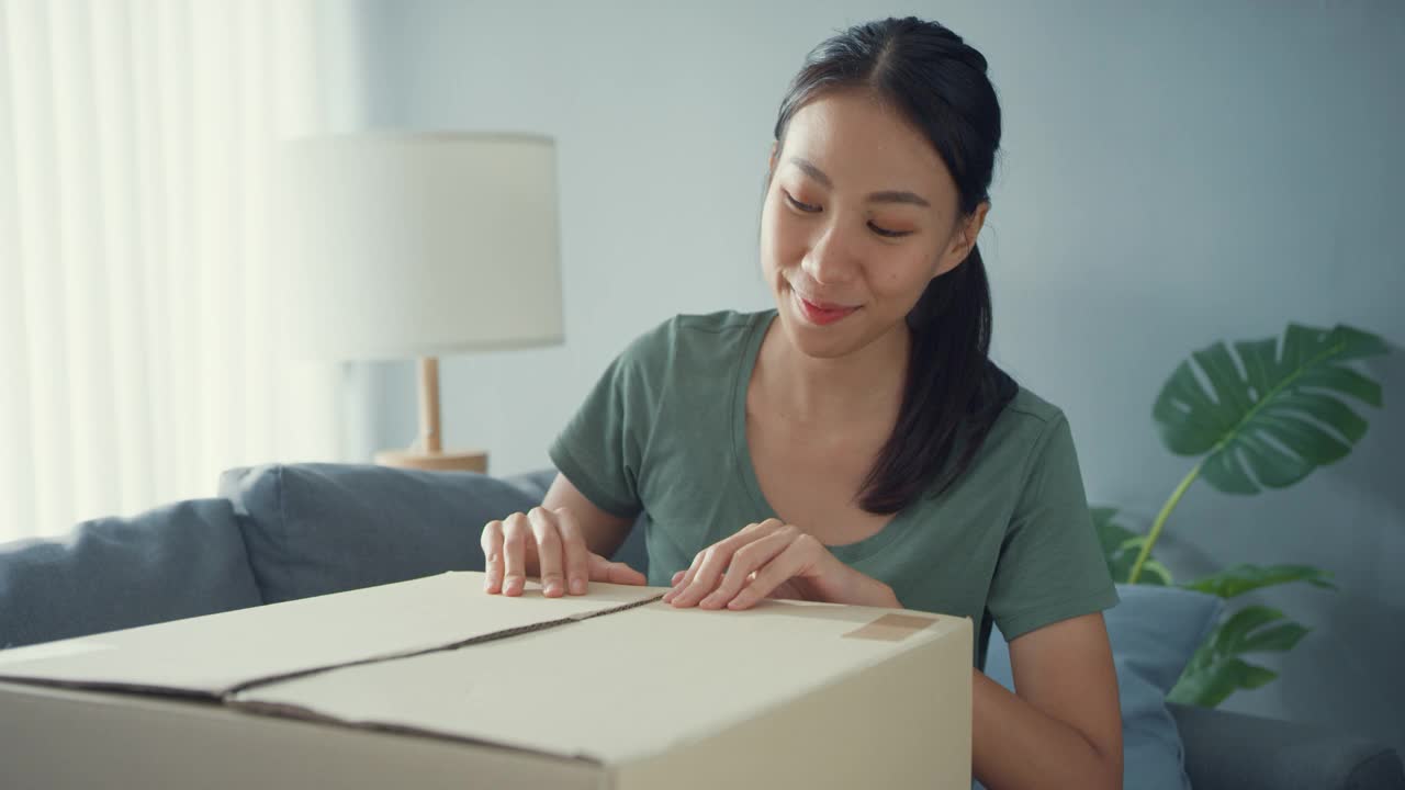 快乐亚洲女士打开纸板箱包装，享受在家中客厅尝试和搭配网上市场的时尚布艺产品。视频素材