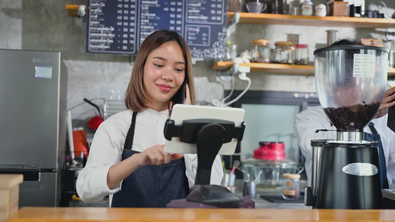 拥有咖啡店和网上订购概念。身穿围裙的亚洲年轻女咖啡师在咖啡店用手机和平板电脑与顾客交谈。视频素材