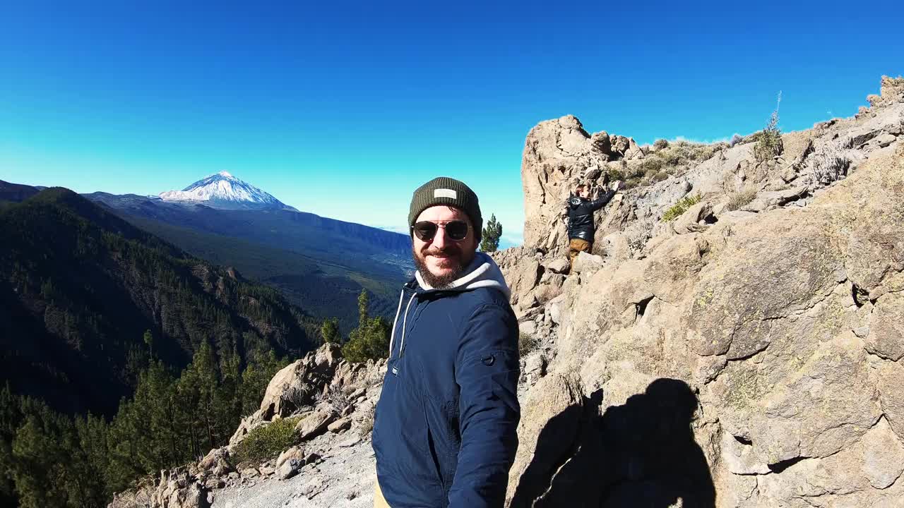 一个戴着太阳镜留着胡子的人爬上了山顶。喜欢欣赏美丽的风景和微笑。女子用智能手机拍照，在山脉和天空的背景下挥手。徒步旅行。度蜜月视频素材