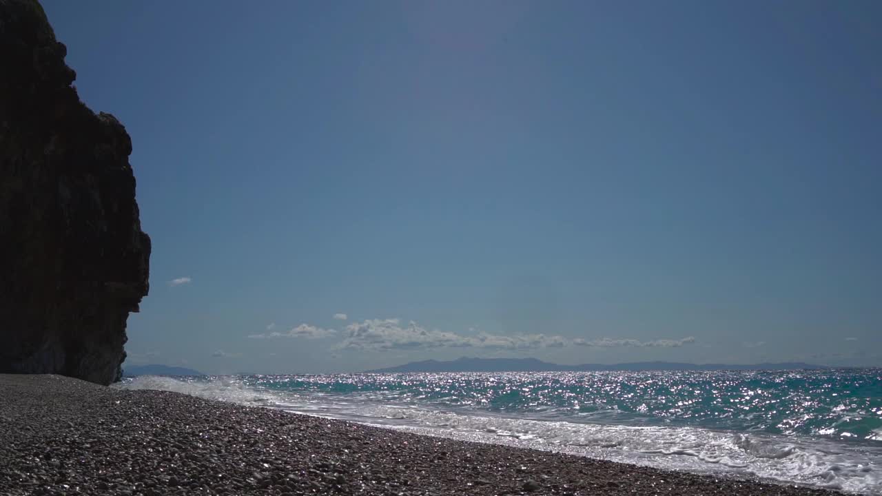 阳光灿烂的日子里，石海岸边的海浪拍打着沙滩，形成白色的泡沫。视频素材